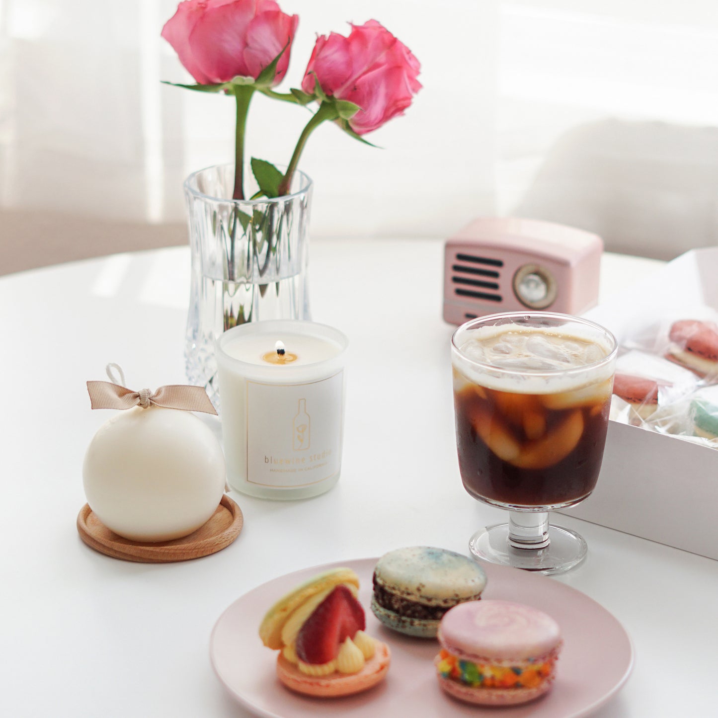bluewine studio lit 5oz frosted glass container soy candle, a white round sphere candle with beige ribbon on the round light wood coaster, colorful macarons on a pink plate, iced americano in a goblet glass, two pink roses in a glass vase, pink speaker, and a box of macarons on the white round table