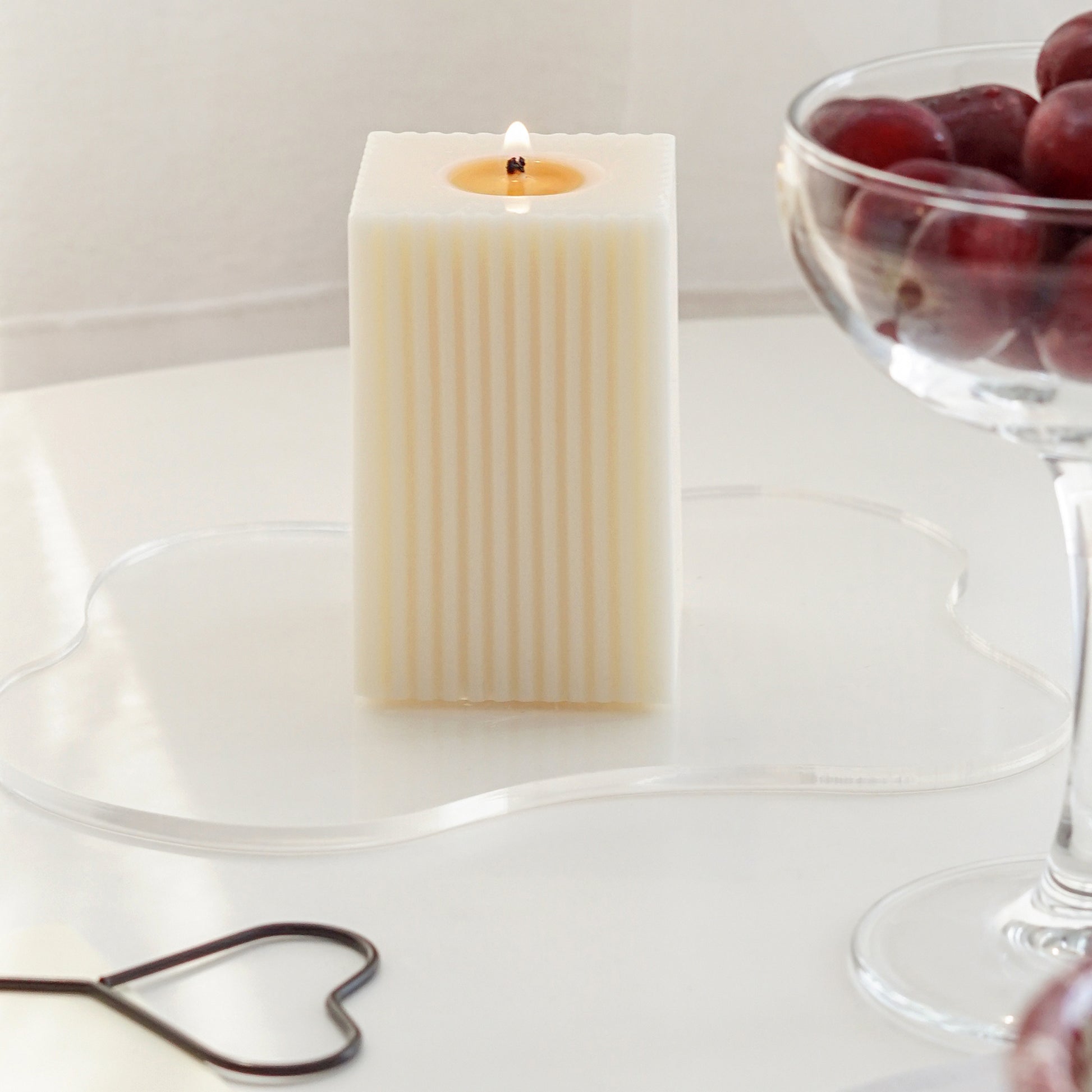 lit square ribbed pillar soy candle on a clear wavy acrylic coaster, a heart shaped candle wick dipper, and a clear coupe glass filled with red cherries on the white round table