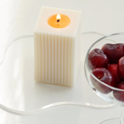 lit square ribbed pillar soy candle on a clear wavy acrylic coaster with a coupe glass filled with red cherries on the white table