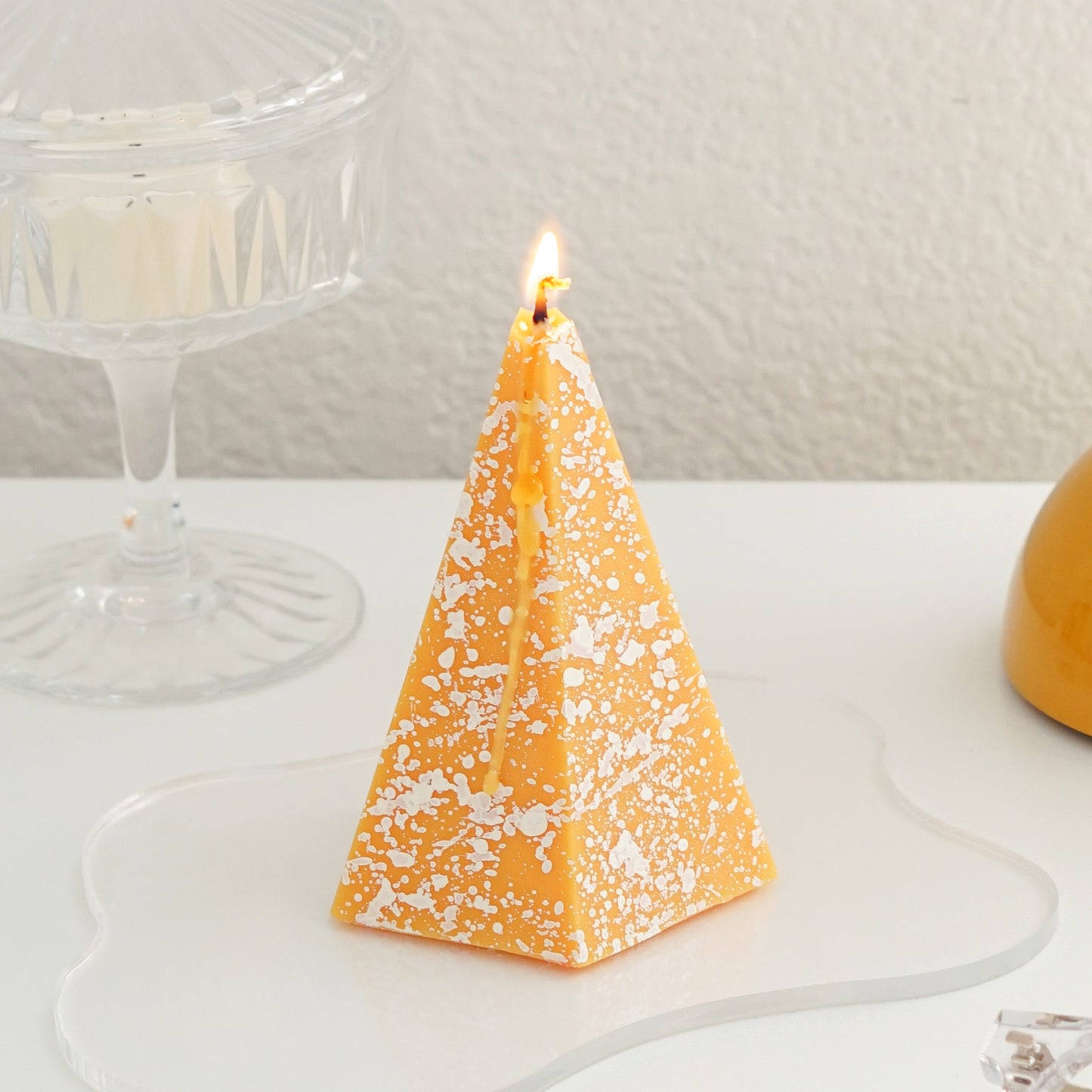 lit pentagonal pyramid shape yellow soy pillar candle with white splash pattern is placed on the white table with ikea clear glass tealight candle holder, yellow &tradition flowerpot lamp, and midnight blue iphone 13
