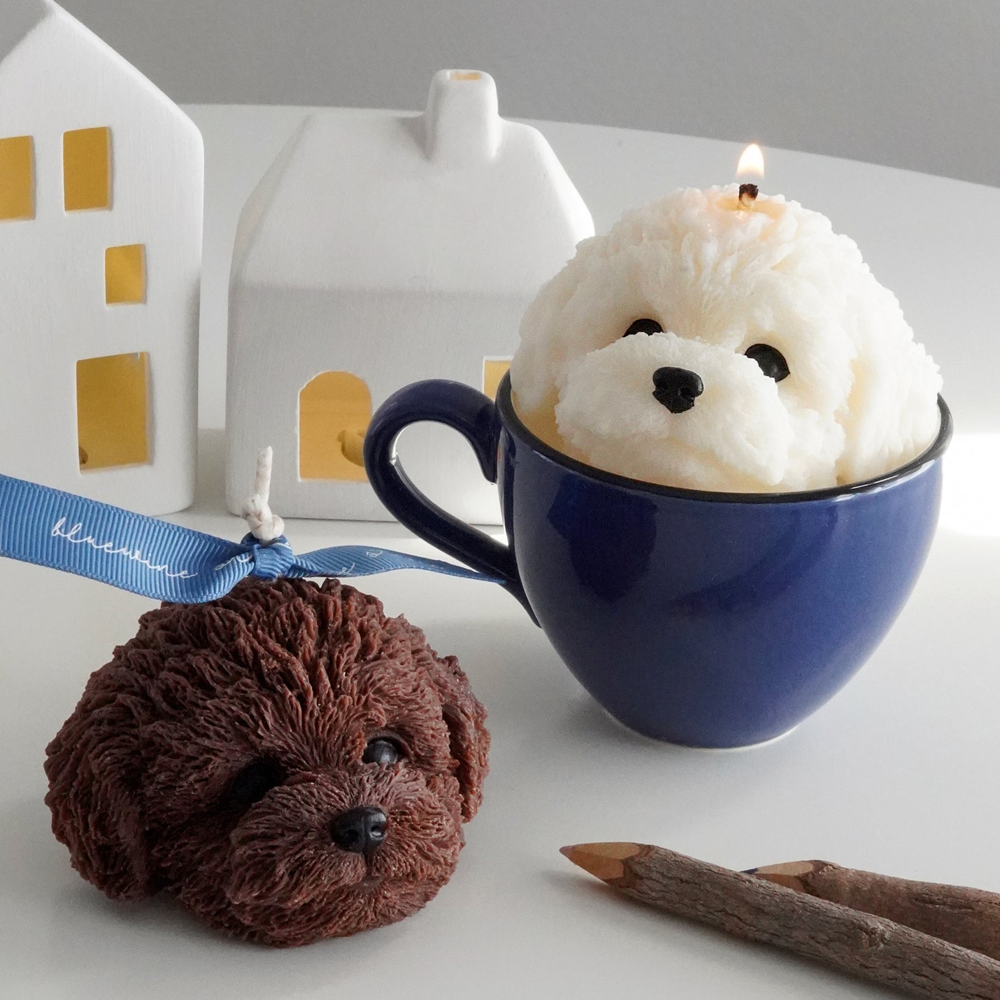 chocolate poodle soy pillar candle with blue bluewine ribbon, a lit white bichon soy pillar candle in a blue mug, wood color pencils, and two house lamps on the white round table