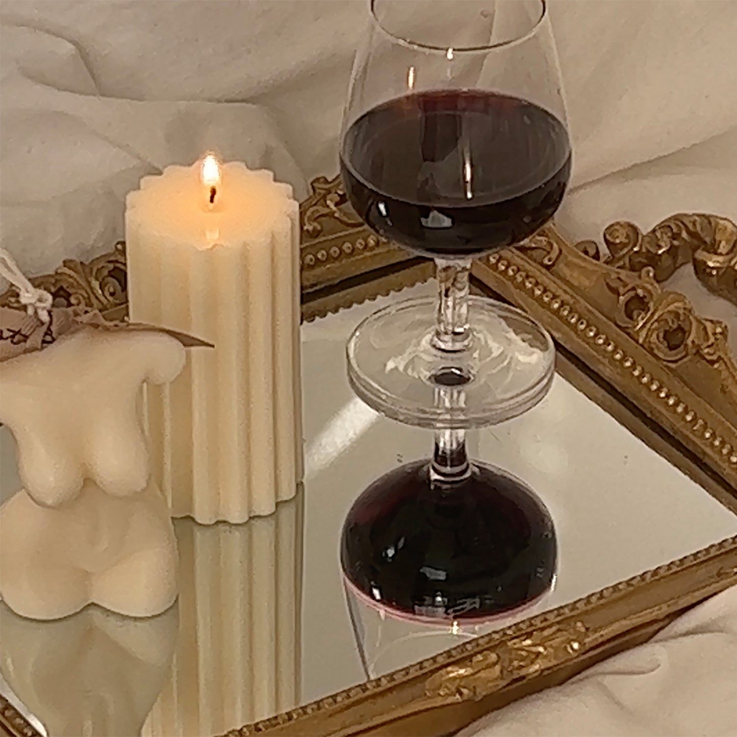 feminine small size body shape white soy pillar candle with beige bluewine studio ribbon, white ribbed soy pillar sculptural candle, and a glass of red wine on the gold french mirror tray placed on bed