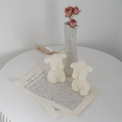 a feminine body shape candle with beige bluewine studio ribbon and another body candle on torn book pages with two matches and pink dried flower roes in a ceramic dotted rough texture unique vase on white round ceramic side table