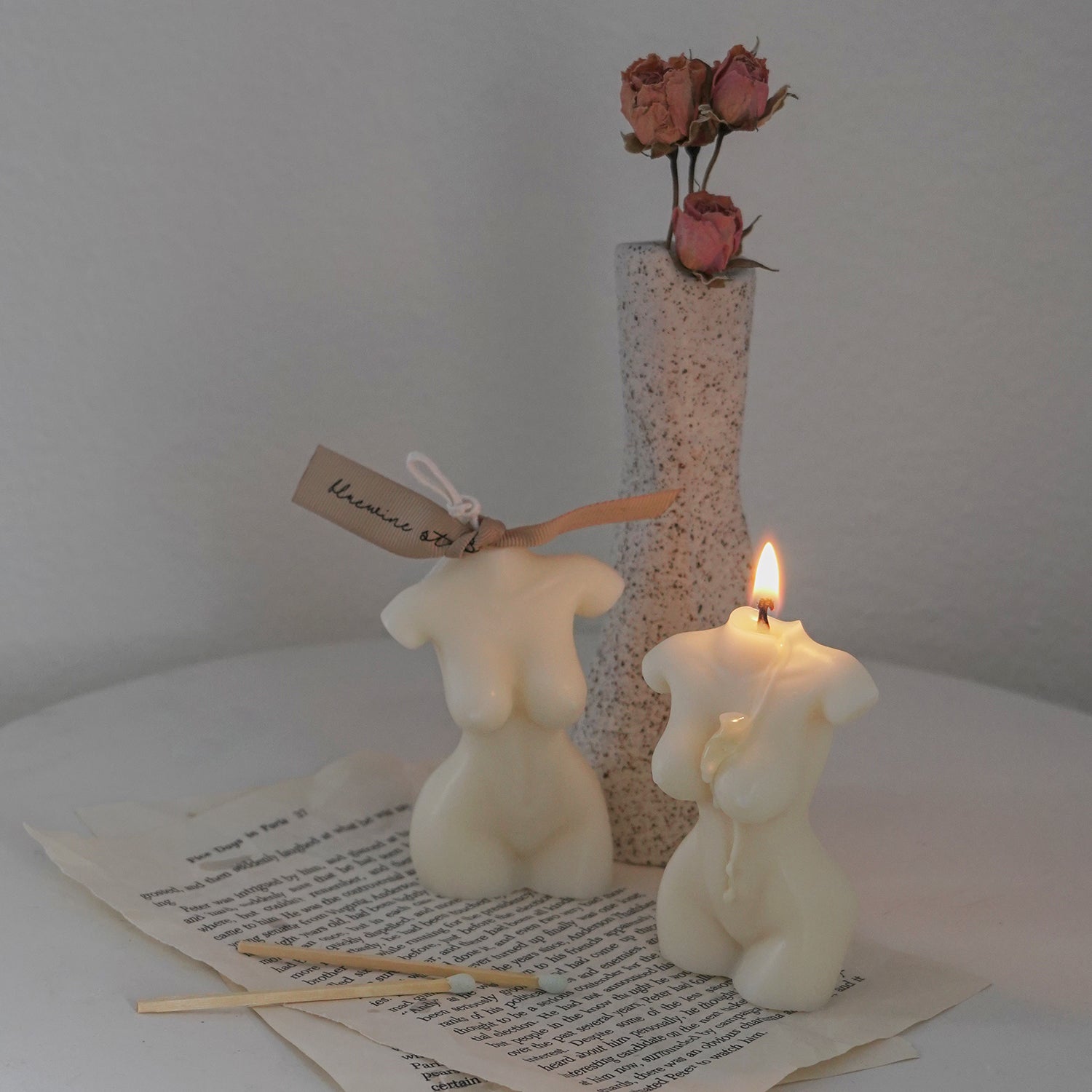 a feminine body shape candle with beige bluewine studio ribbon and a lit body candle on torn book pages with two matches and pink dried flower roes in a ceramic dotted rough texture unique vase on white round ceramic side table