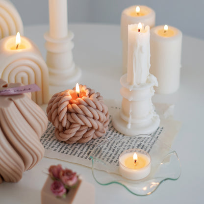 a lit beige yarn ball shape soy pillar candle, a lit tealight candle on a clear wavy ruffle dish, a candlestick shape pillar candle on book pages
