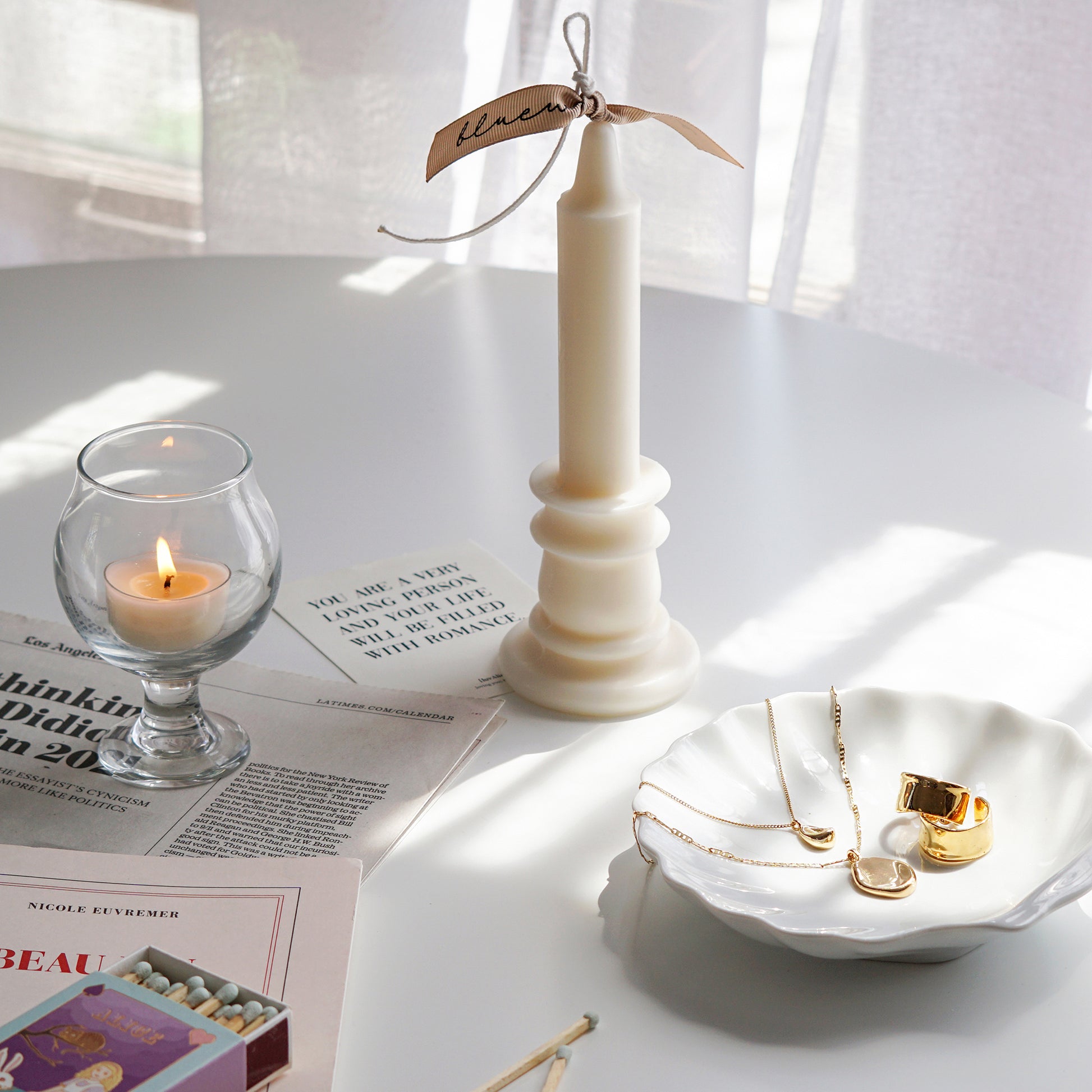 candlestick shape white soy pillar candle with beige bluewine studio ribbon, a lit tealight candle in a mini glass, mint color matches, newspapers, a postcard, French book called Beau Jeu, gold earrings and necklace on white ceramic shell tray on white round table
