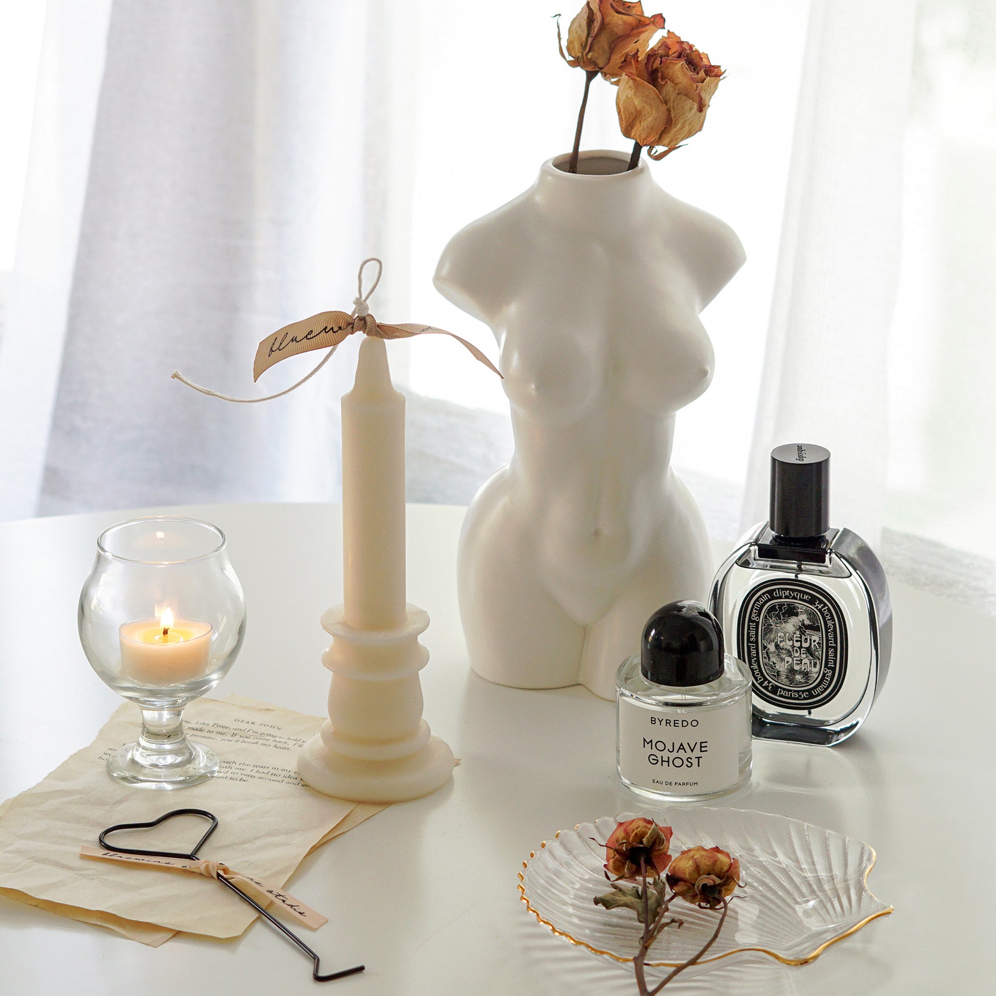 white candlestick shape soy pillar candle with beige bluewine studio ribbon, a lit tealight candle in a mini glass, and a heart wick dipper on book pages, orange dried roses on gold rim clear shell tray, Byredo Mojave Ghost, Diptyque Fleur de Peau, and feminine body vase with dried roses on white round table