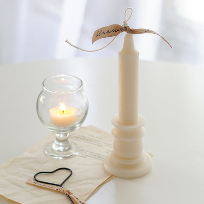 white candlestick shape soy pillar candle with beige bluewine studio ribbon, heart wick dipper, book pages, and a lit tealight candle in a mini glass on white round table