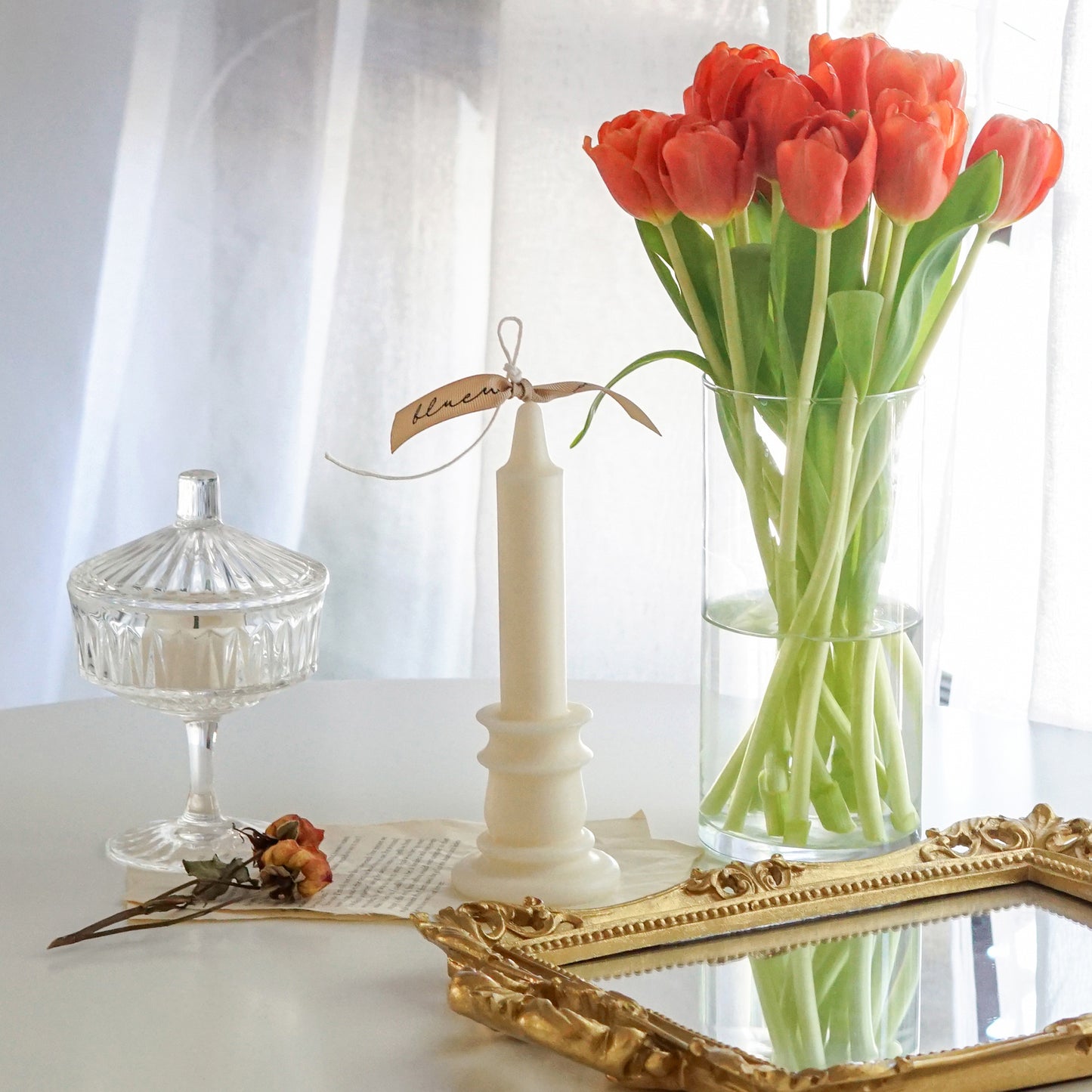 candlestick shape white soy pillar candle with beige bluewine studio ribbon, coupe glass and orange dried roses on book pages, rectangular gold french mirror tray, and orange tulips in a clear round vase on white round table