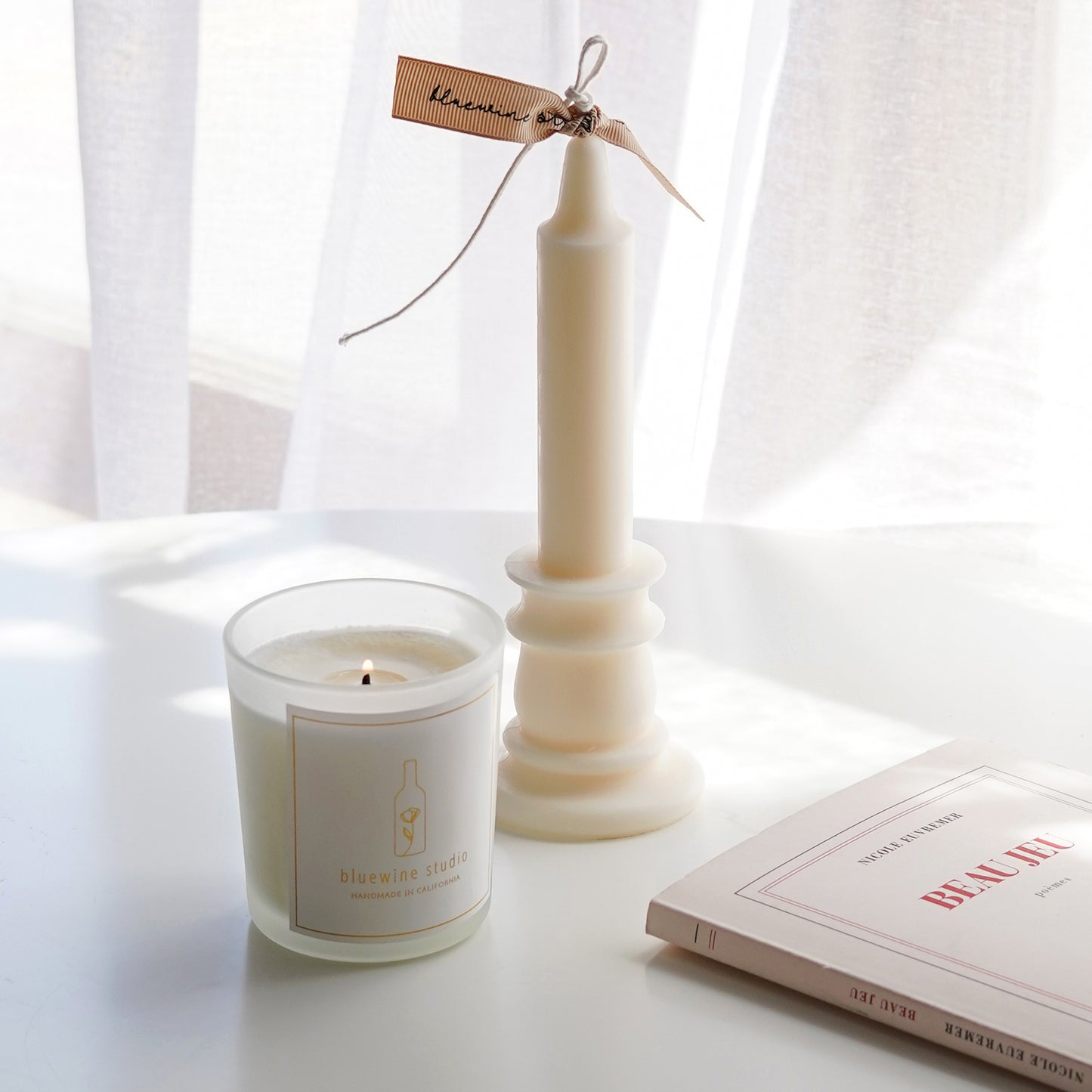 bluewine studio lit 5oz frosted glass container candle, a white candlestick shape candle with a beige ribbon, and french book Beau Jeu on the white round table
