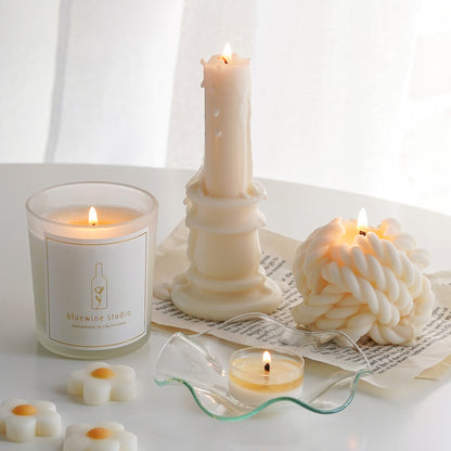 bluewine studio lit 5oz soy container candle, a white melted candlestick candle and a yarn candle on the book papers, a tealight candle on a clear ruffle dish, and daisy wax melts on the white round table