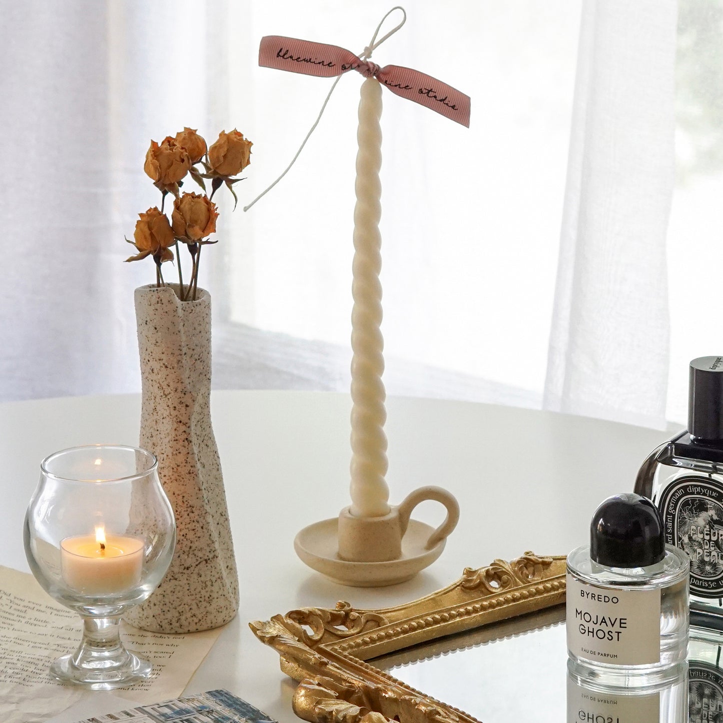 white twisted swirl soy pillar taper candle in a dotted beige ceramic candle holder, yellow rose dried flowers in a dotted ceramic unique vase and a lit tealight candle in a mini glass placed on book pages, and Byredo Mojave Ghost and Diptyque Fleur de Peau placed on rectangular gold french mirror tray on white round table