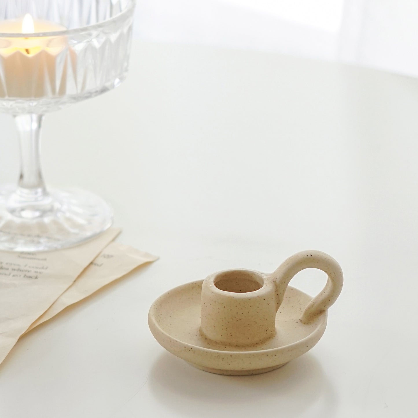 a dotted neutral beige color ceramic taper candle holder and a lit tealight candle in a coupe glass placed on book pages on white round table