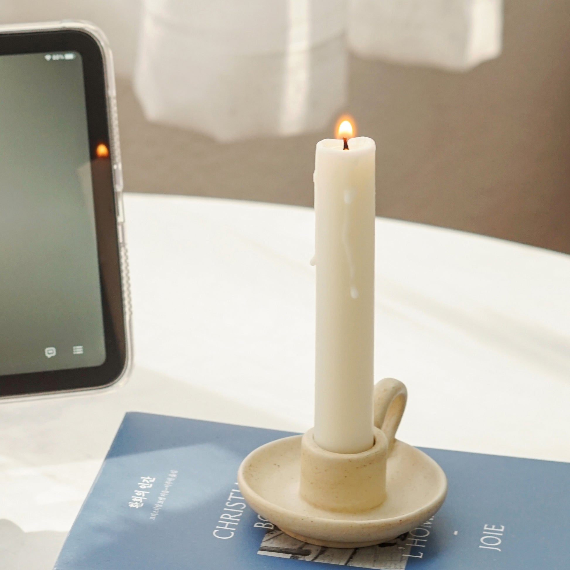 a lit white soy pillar taper candle in a beige neutral color candle holder placed on blue book and ipad mini playing the song Japanese Denim by Daniel Ceaser