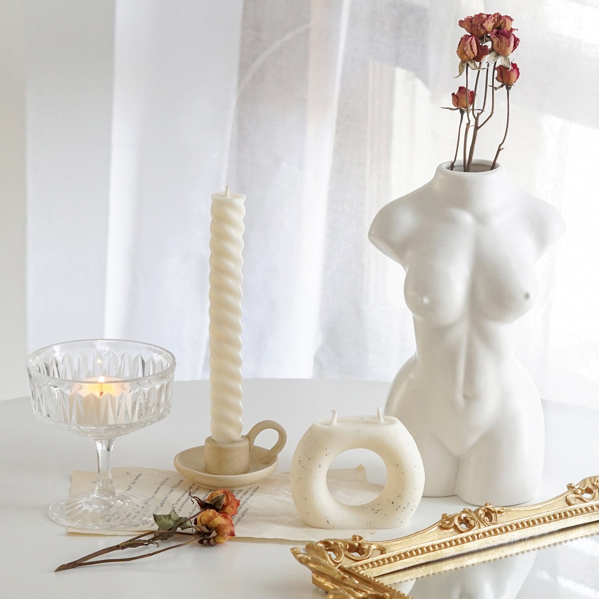 white twisted swirl taper candle in a ceramic candle holder, orange rose dried flowers, ring donut shape dotted white soy pillar candle, a lit tealight candle in a coupe glass placed on book pages, rectangular gold french mirror tray, and a feminine body shape white ceramic flower vase on white round table