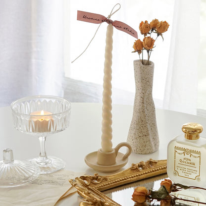 white twisted soy pillar candle with pink bluewine studio ribbon in a dotted beige ceramic taper candle holder, a lit tealight candle in a coupe glass placed on book pages, and yellow rose dried flowers and santa maria novella perfume placed on rectangular gold french mirror tray on white table