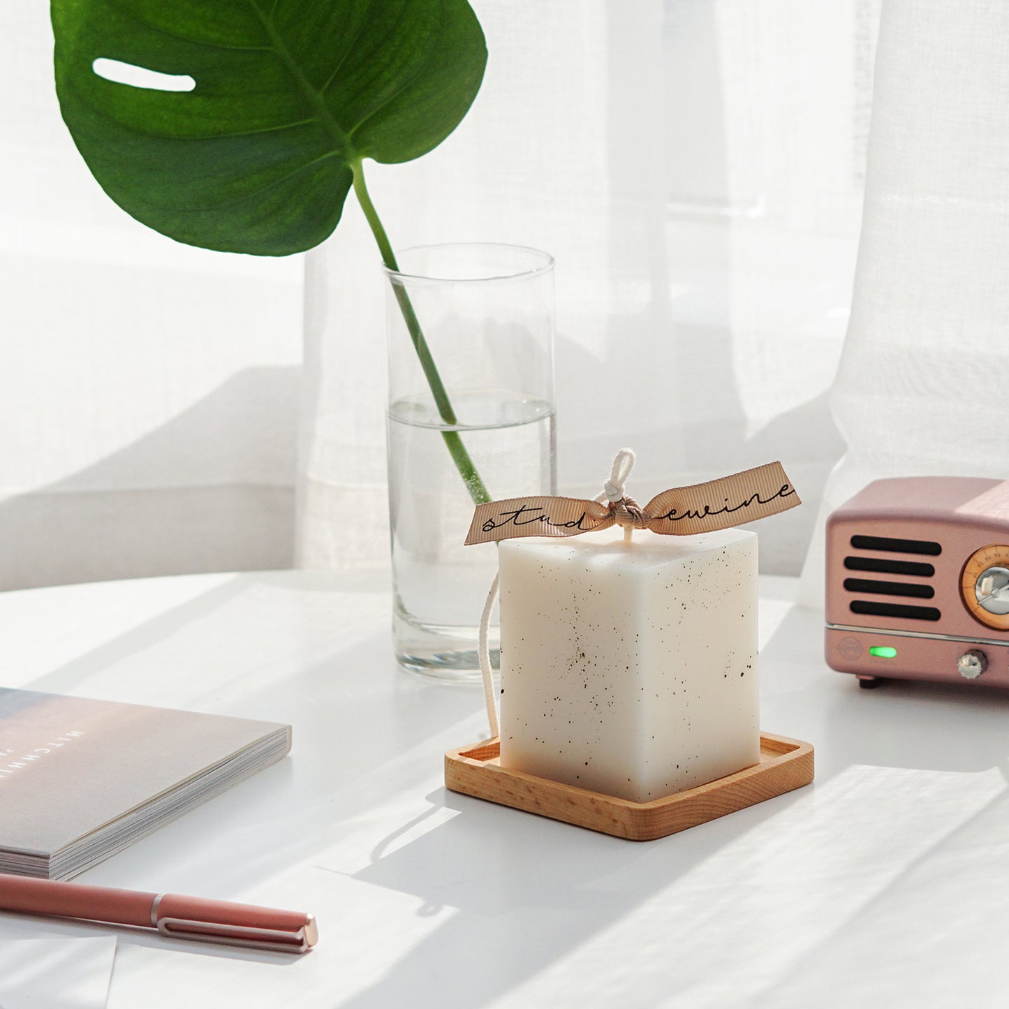 a dotted white square pillar candle with beige bluewine studio ribbon on a square wood coaster, a single monstera leaf in a glass vase, a book, a pink pen, and a pink speaker on white round table