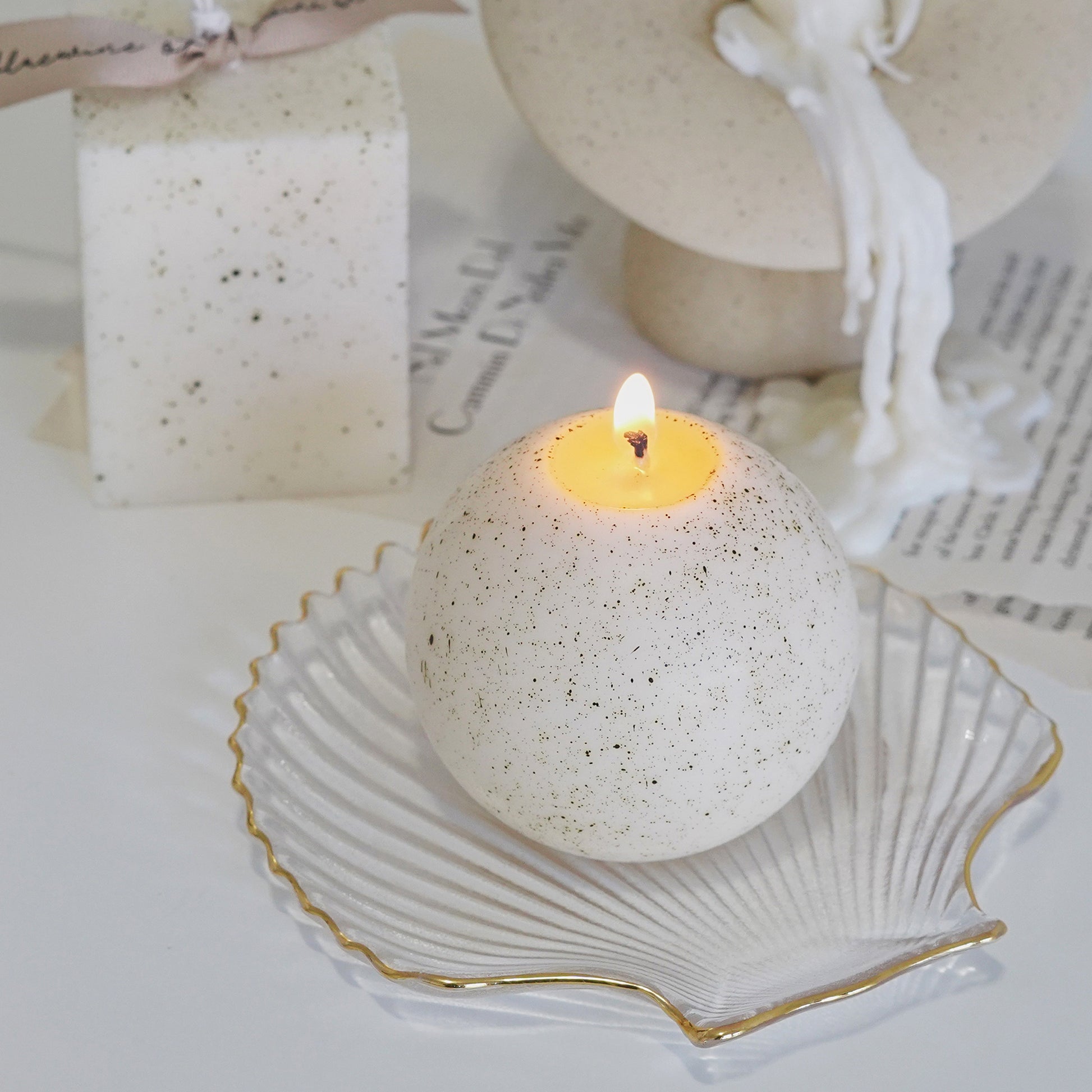 a lit dotted white sphere ceramic pattern soy pillar candle on a gold rim clear seashell tray and a dotted white square pillar candle with beige bluewine studio ribbon