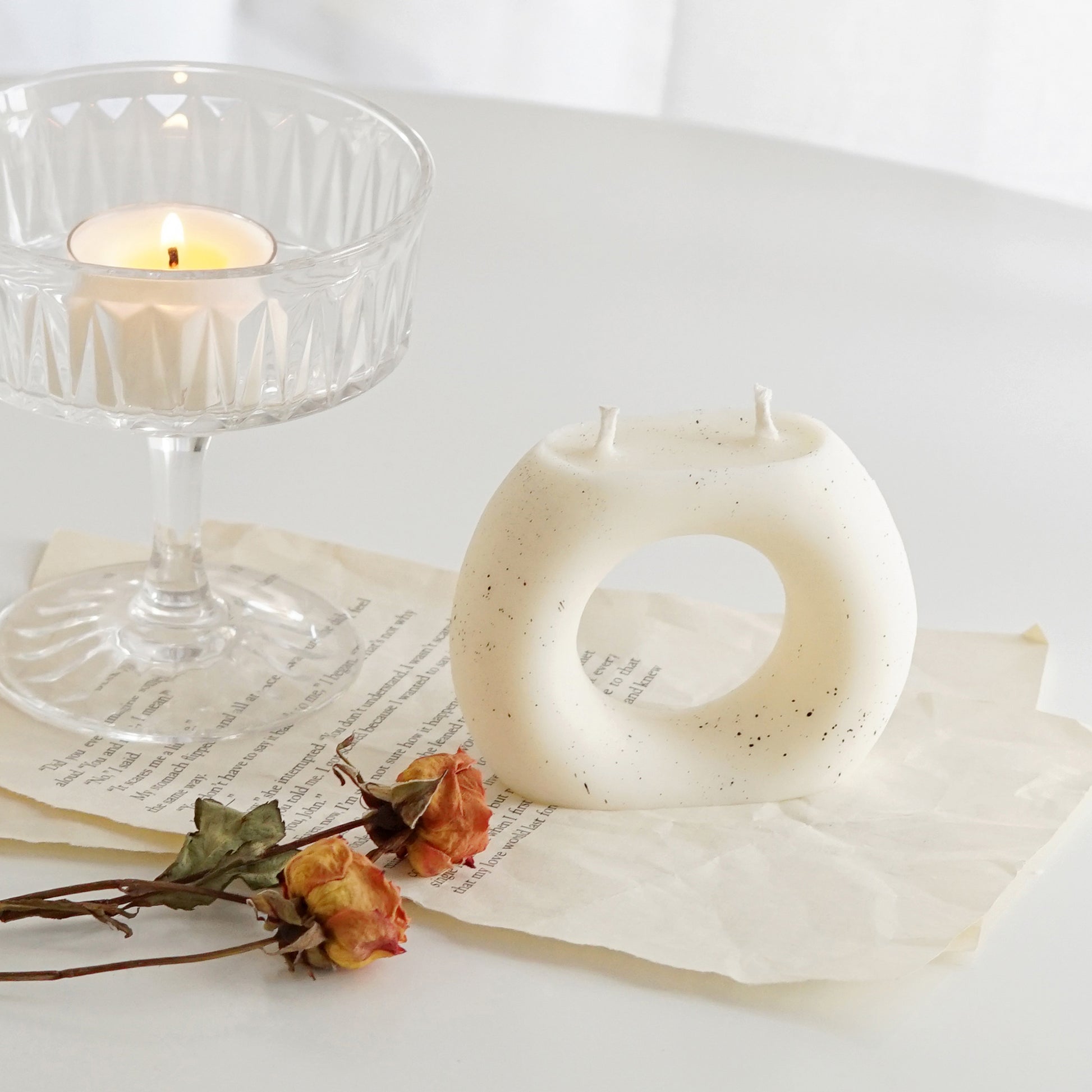a dotted ring donut shaped white soy pillar candle, yellow orange rose dried flowers, and a lit tealight candle in a glass coupe placed on book pages on white round table