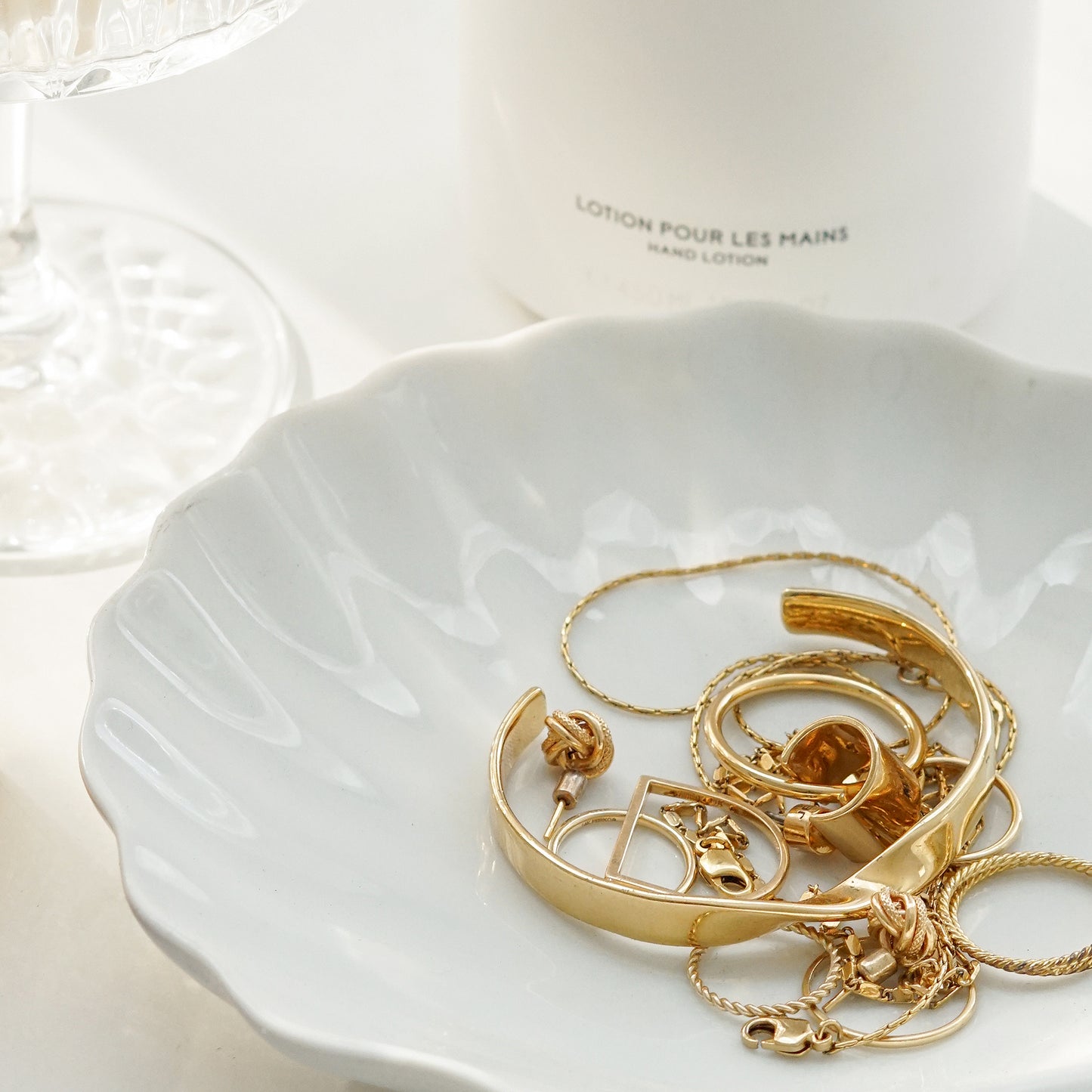 coupe glass, Byredo body lotion, and gold bracelet, earrings, and rings on ceramic shell tray