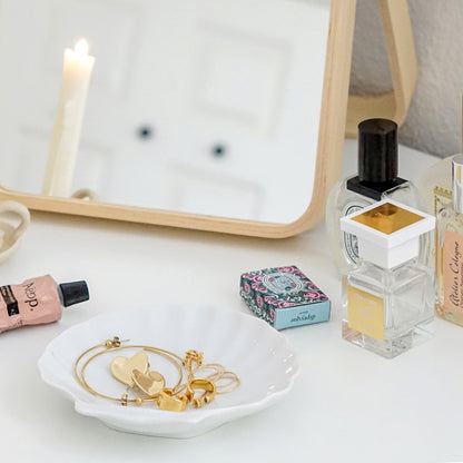 a lit white soy pillar taper candle in a ceramic candle holder, Aesop hand lotion, gold earrings and rings on ceramic shell ray, Tom Ford White Suede, Atelier Cologne Orange Sanguine, Diptyque Rose, Santa Maria Novella perfume, Ikea vanity mirror placed on white Ikea closet