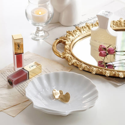 Jenny Bird Heart shape earrings on white ceramic shell tray and ysl lip tints on book pages, a lit tealight candle in a mini glass, pink rose dried flowers and Creed perfume on round gold french mirror tray