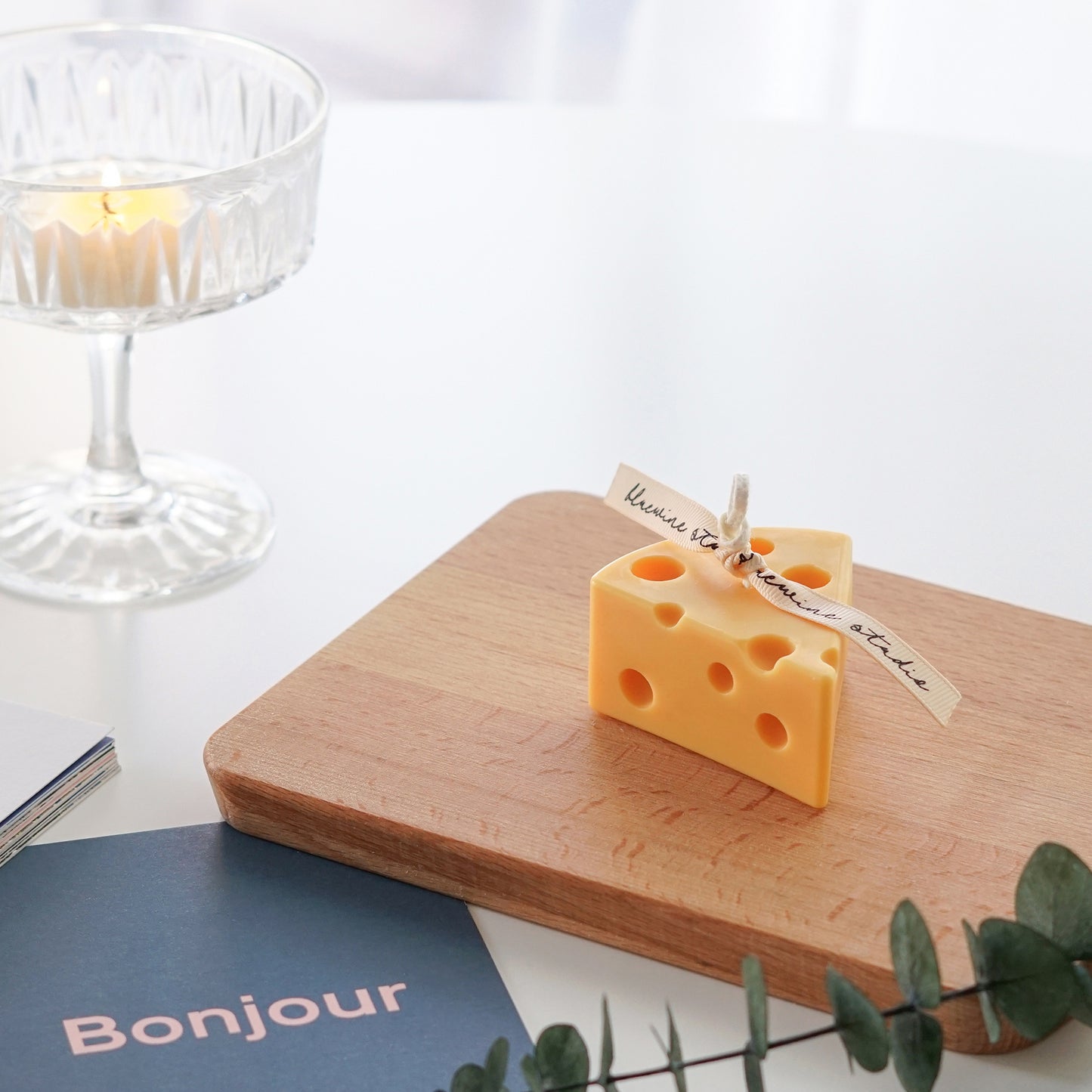 a slice of cheese soy pillar candle on wood board, eucalpytus, a postcard, and a lit tealight candle in a cocktail glass