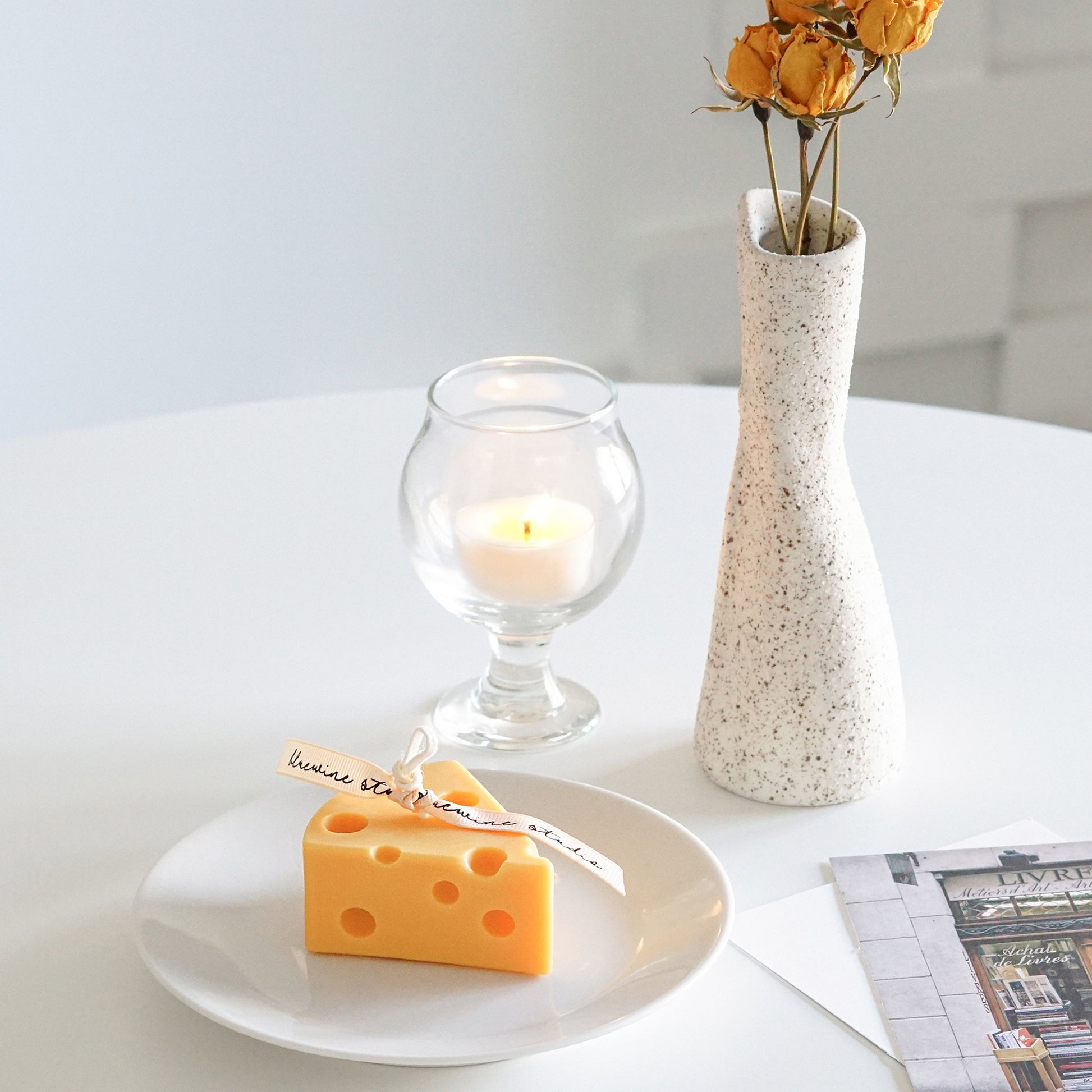 slice of cheese soy pillar candle on white dish, a lit tealight candle in a mini glass, yellow dried roses in a ceramic vase, and postcards on white round table