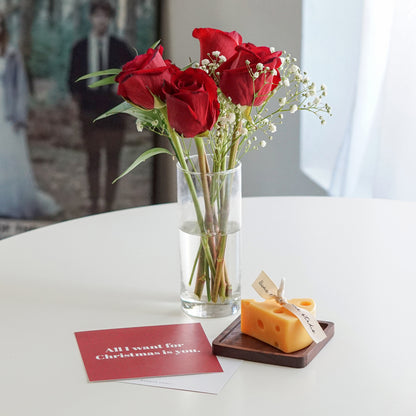 a slice of cheese soy pillar candle on a wood coaster, christmas postcards, and roses in a clear in vase on white round table