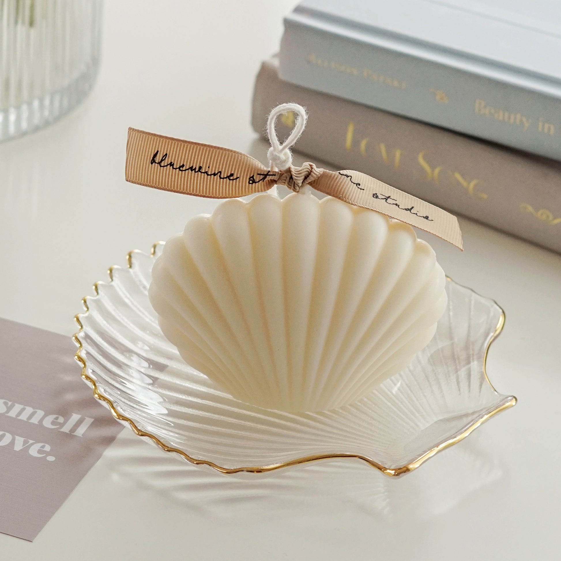 white seashell soy pillar candle with beige bluewine studio ribbon on a gold rim clear shell tray, two books, you smell like love beige postcard