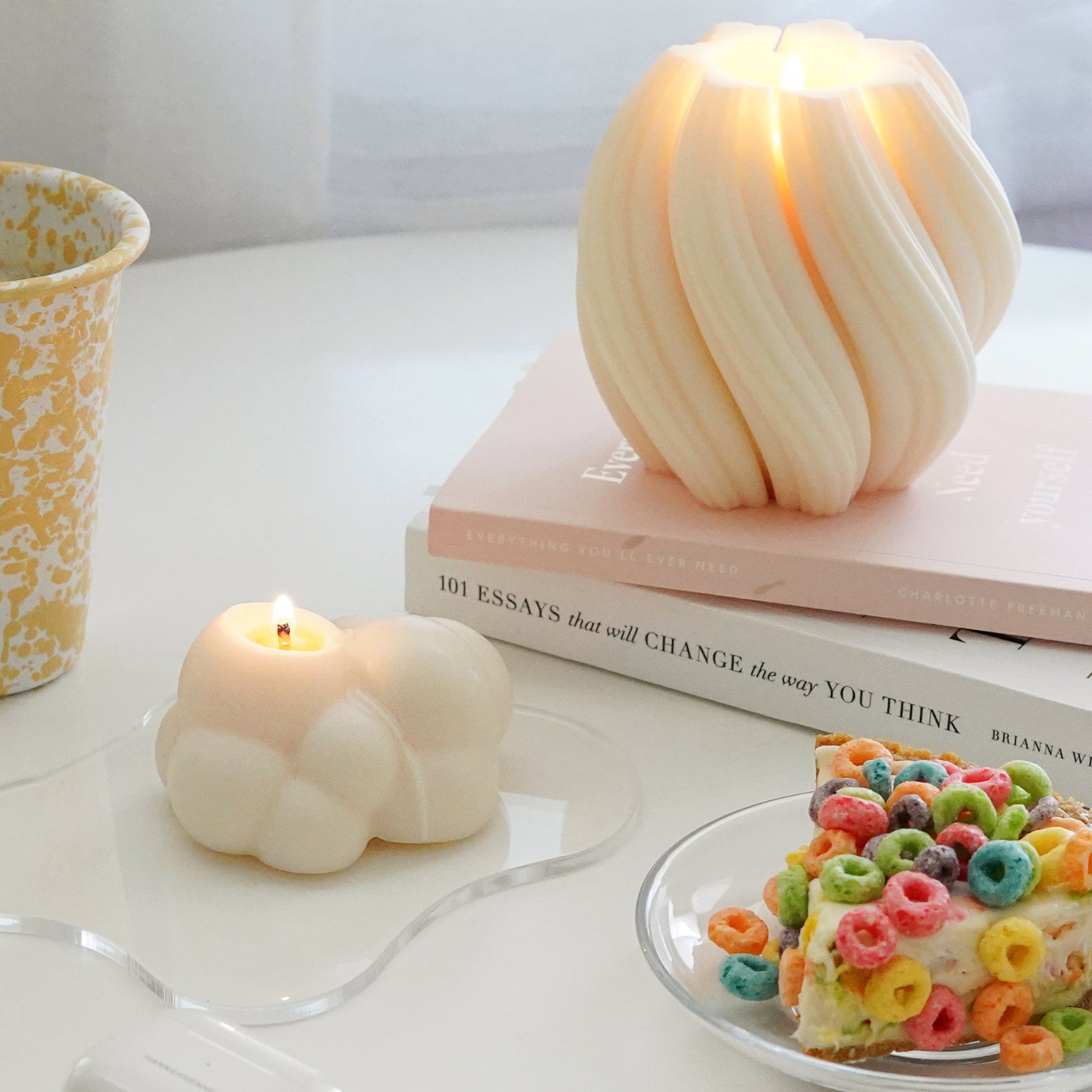 a lit dreamy aesthetic white cloud soy pillar candle on a clear wavy acrylic coaster and a cereal pie from pie hole on a clear dish, yellow crow canyon tumbler mug, and a lit sculptural minimal swirl objet candle on two books