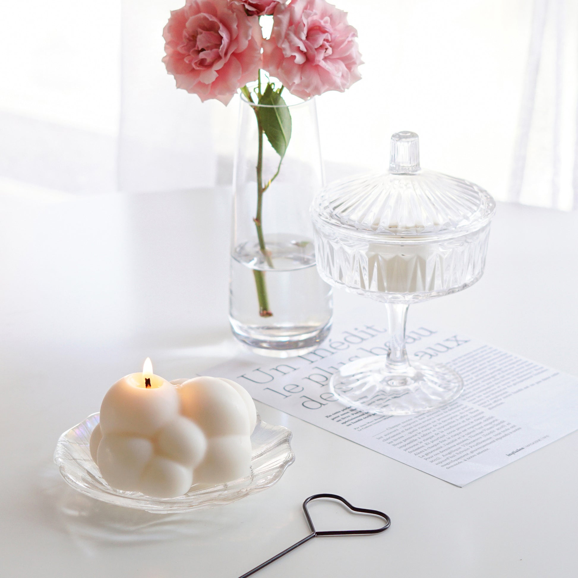a lit white cloud soy pillar candle on a holographic shell tray, heart wick dipper, and a magazine paper, a lit tealight candle in a mini glass, and pink flowers in a clear cylinder vase