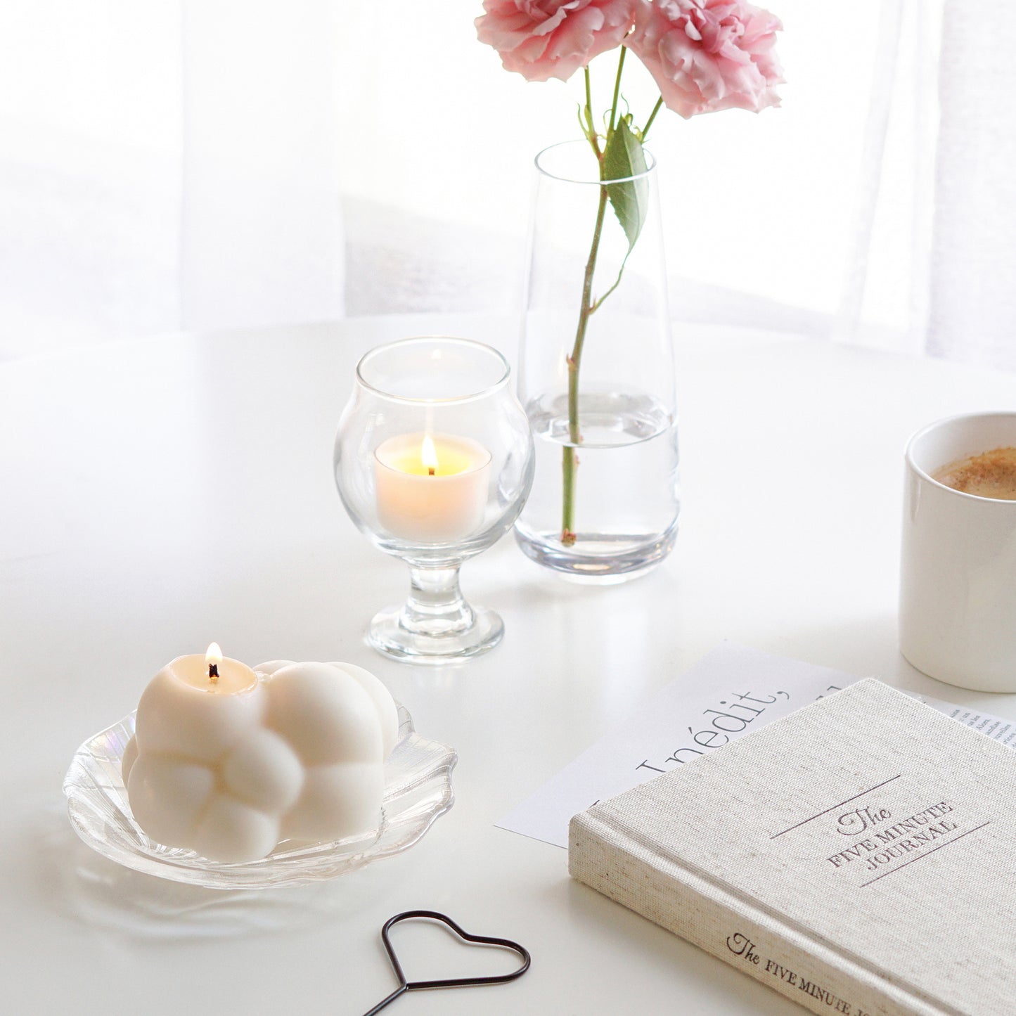 a lit white cloud soy pillar candle on a holographic shell tray, heart wick dipper, the five minute journal, and a magazine paper, a lit tealight candle in a mini glass, hot coffee in a white mug, and pink flowers in a clear cylinder vase