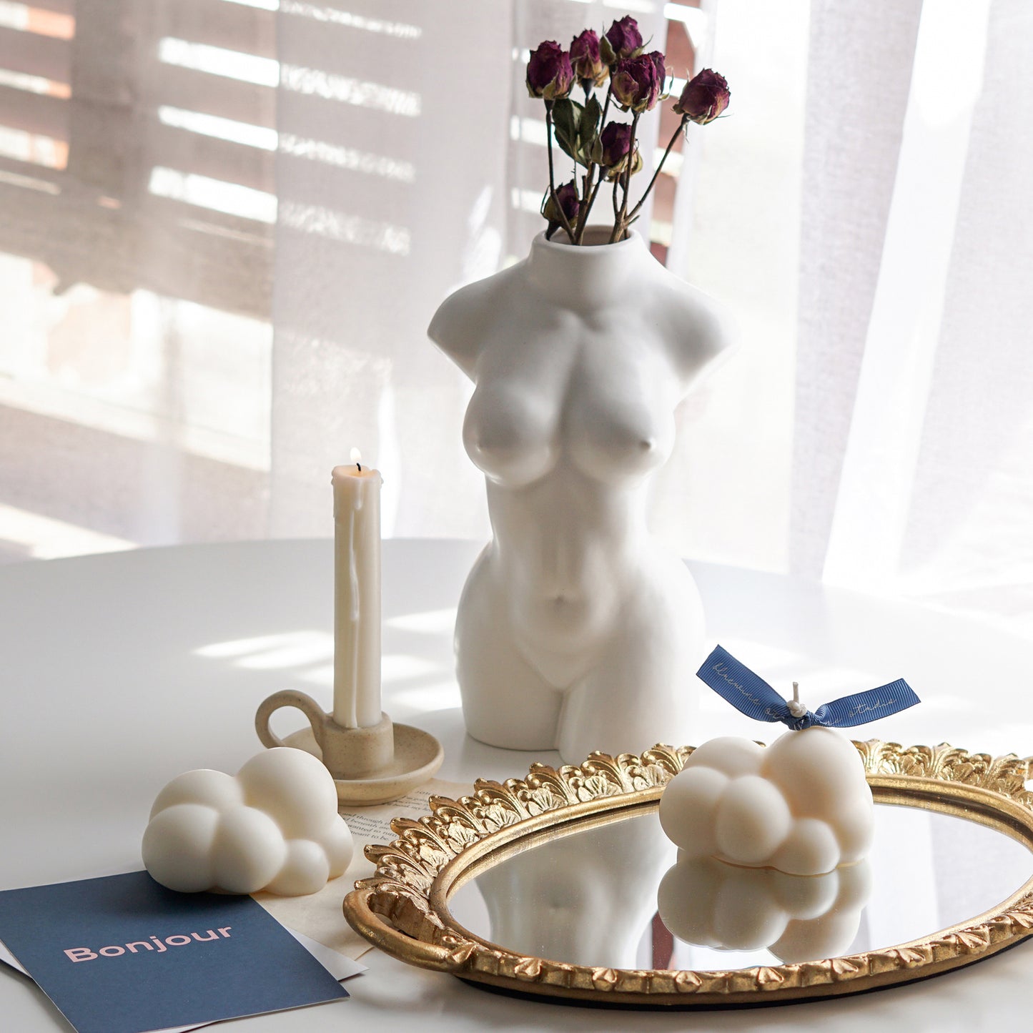 white cloud soy pillar candle with a blue bluewine studio ribbon on a round gold french mirror tray, cloud wax melt, blue bonjour postcard, a lit taper candle in a ceramic candle holder, and burgundy dried flowers in a ceramic feminine body vase
