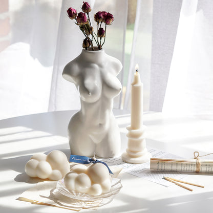 white cloud soy pillar candle on a holographic shell tray, a cloud wax melt, rolled book pages, matches, and a lit candlestick shape soy pillar candle, and burgundy dried roses in a white ceramic feminine body vase