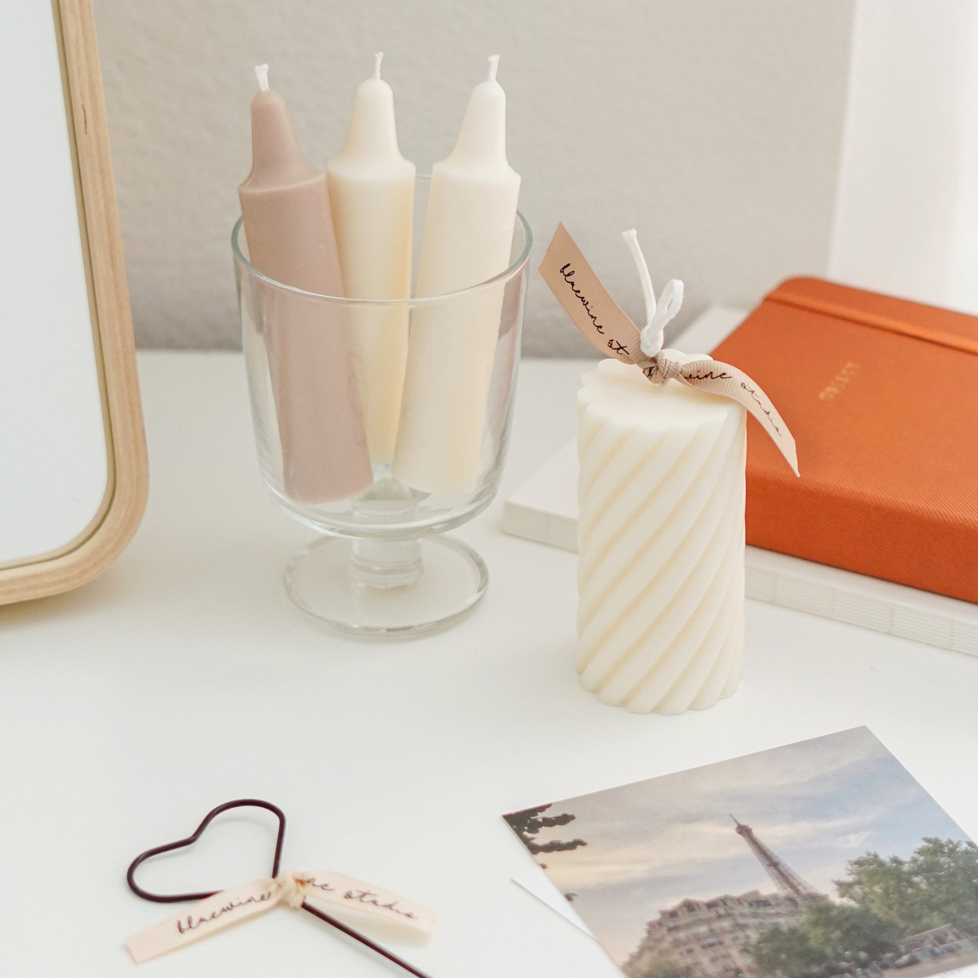 three crayon shape soy pillar candles in a glass, heart wick dipper, hermes perfume, aesop hand lotion, eiffel tower postcards, a spiral white soy pillar candle with a beige bluewine studio ribbon, orange planner, and ikea wood vanity mirror