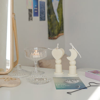 Heart shape candle, crescent moon shape candles, a lit tealight candle in a glass coupe, blue postcard, match box, black heart shape wick dipper, and silver rings on clear beaded tray are place on white drawer. The arrangement exudes minimal aesthetic dreamy vibes.