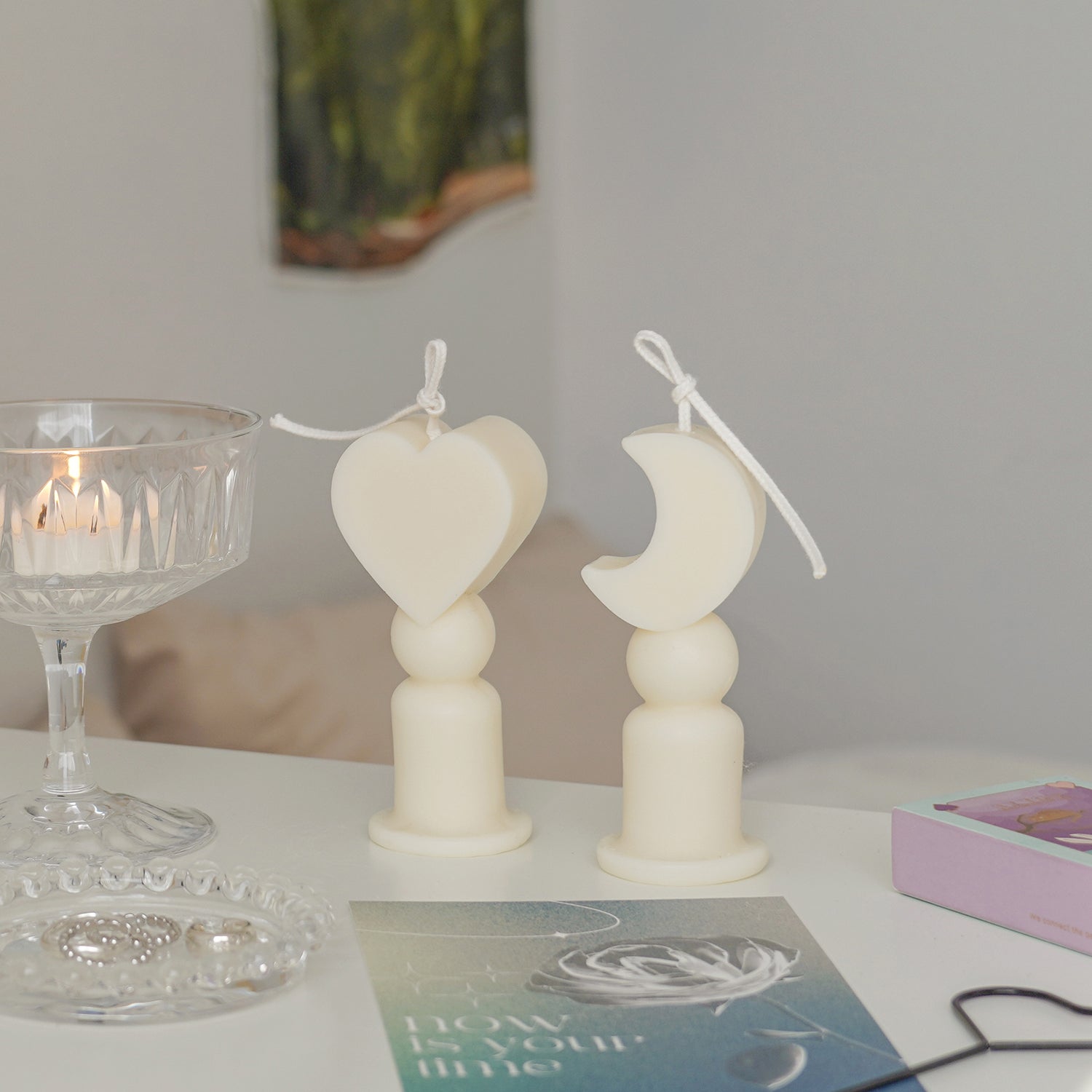 Heart shape candle, crescent moon shape candles, a lit tealight candle in a glass coupe, blue postcard, match box, black heart shape wick dipper, and silver rings on clear beaded tray are place on white drawer. The arrangement exudes minimal aesthetic dreamy vibes.