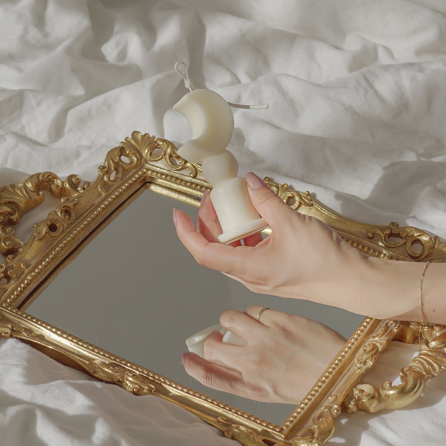 French style rectangular shape mirror placed on a white sheet of bed and a hand holding a crescent moon shape candle.