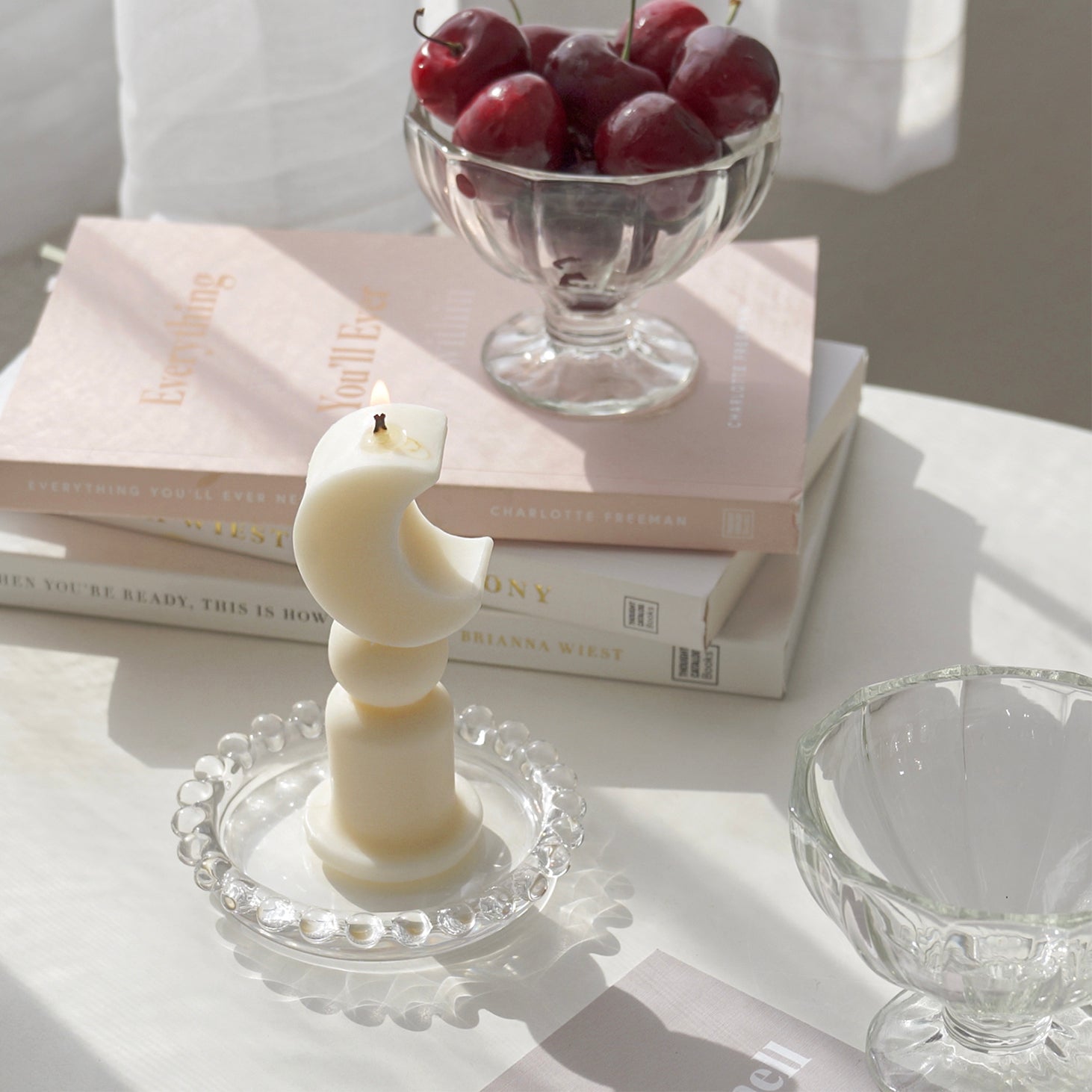 a lit crescent moon shape candles and cherries in a clear ice cream bowl on stacked books are placed on white table. The arrangement exudes minimal aesthetic dreamy vibes.