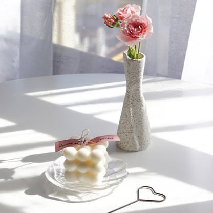 a white bubble soy pillar candle on holographic shell tray, black heart wick dipper, and pink flowers in ceramic vase on white table