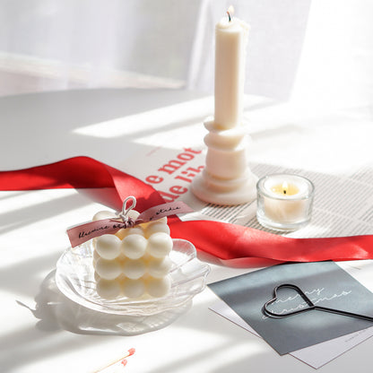 a white square cube bubble soy pillar candle with pink bluewine studio ribbon on a holographic shell tray and merry christmas hunter green postcard, heart wick dipper, red ribbon, a lit tealight candle in a glass tealight candle holder, a lit candlestick shape pillar candle, and magazine paper on white round table