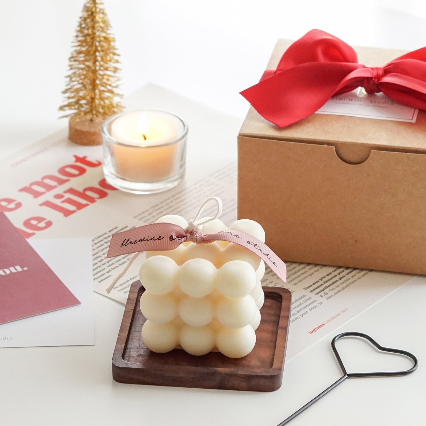 a cube shape soy pillar candle on rectangular wooden coaster, black heart wick dipper, gift box, tealight candle, and christmas tree decor on white table