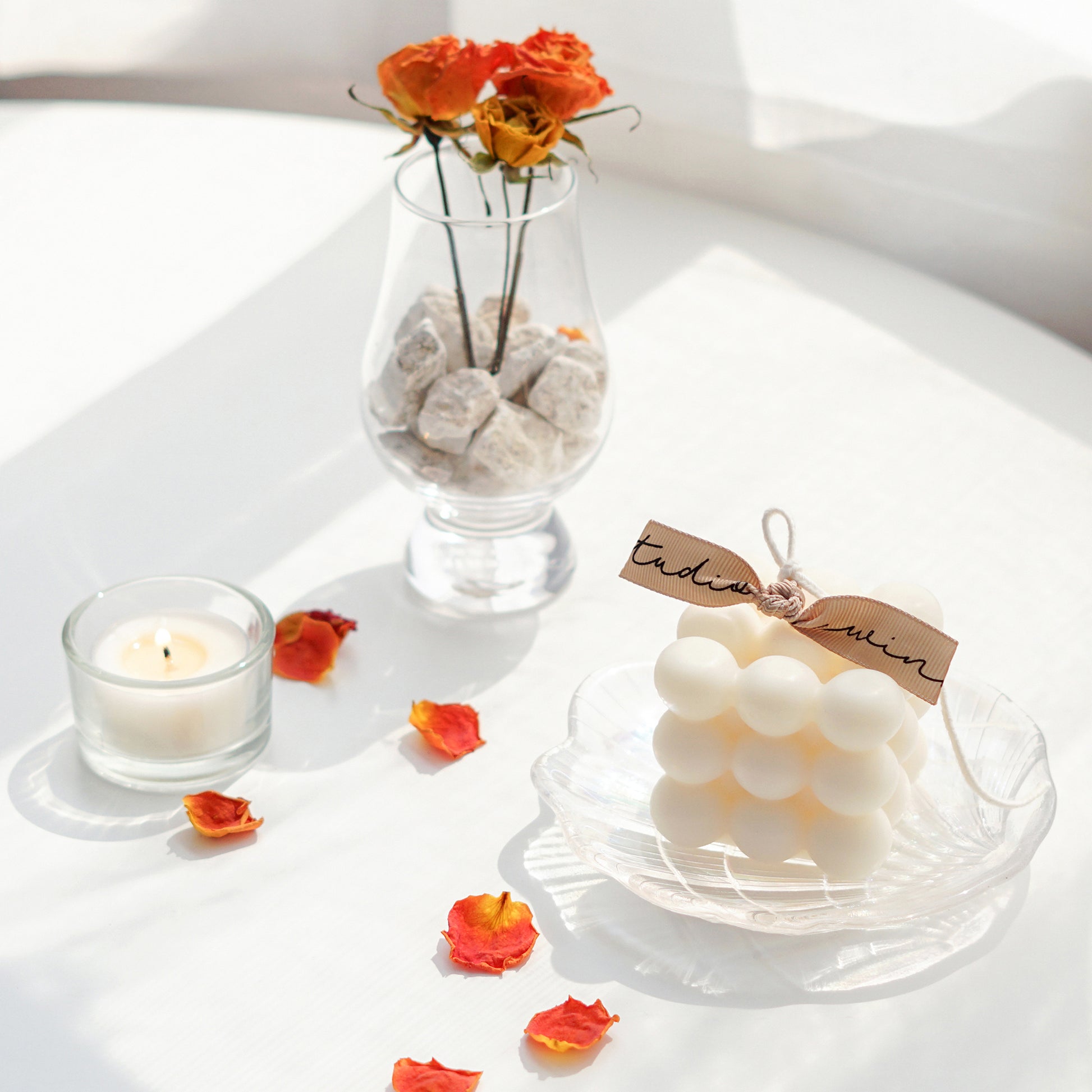 a white cube square bubble soy pillar candle on a holographic shell tray, a lit tealight candle in a tealight glass holder, orange dried roses in a brandy glass with white rocks, and rose petals on white round table