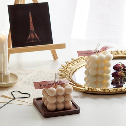 beige square bubble cube soy pillar candle with pink bluewine studio ribbon on a wood square coaster, heart wick dipper, a lit taper candle in a ceramic candle holder, night view Eiffel Tower post card on mini wood easel, white cube candle and burgundy rose dried flowers on gold round oval french mirror tray