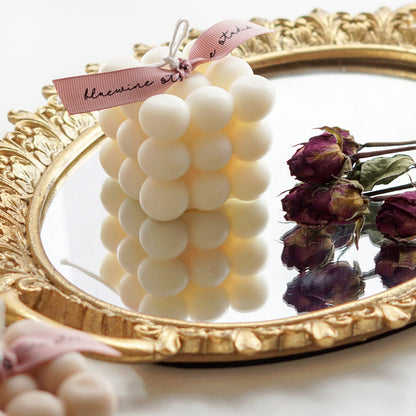 a white square cube bubble soy pillar candle with pink bluewine studio ribbon and burgundy dried roses on a round oval gold french mirror tray