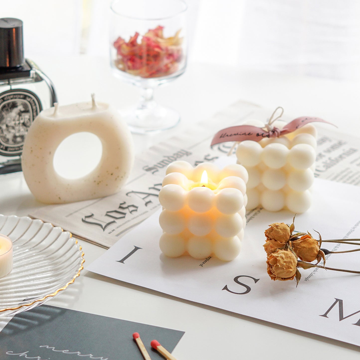 a lit white cube shape bubble soy pillar candle, dried flowers, postcards, newspaper, perfume on white table