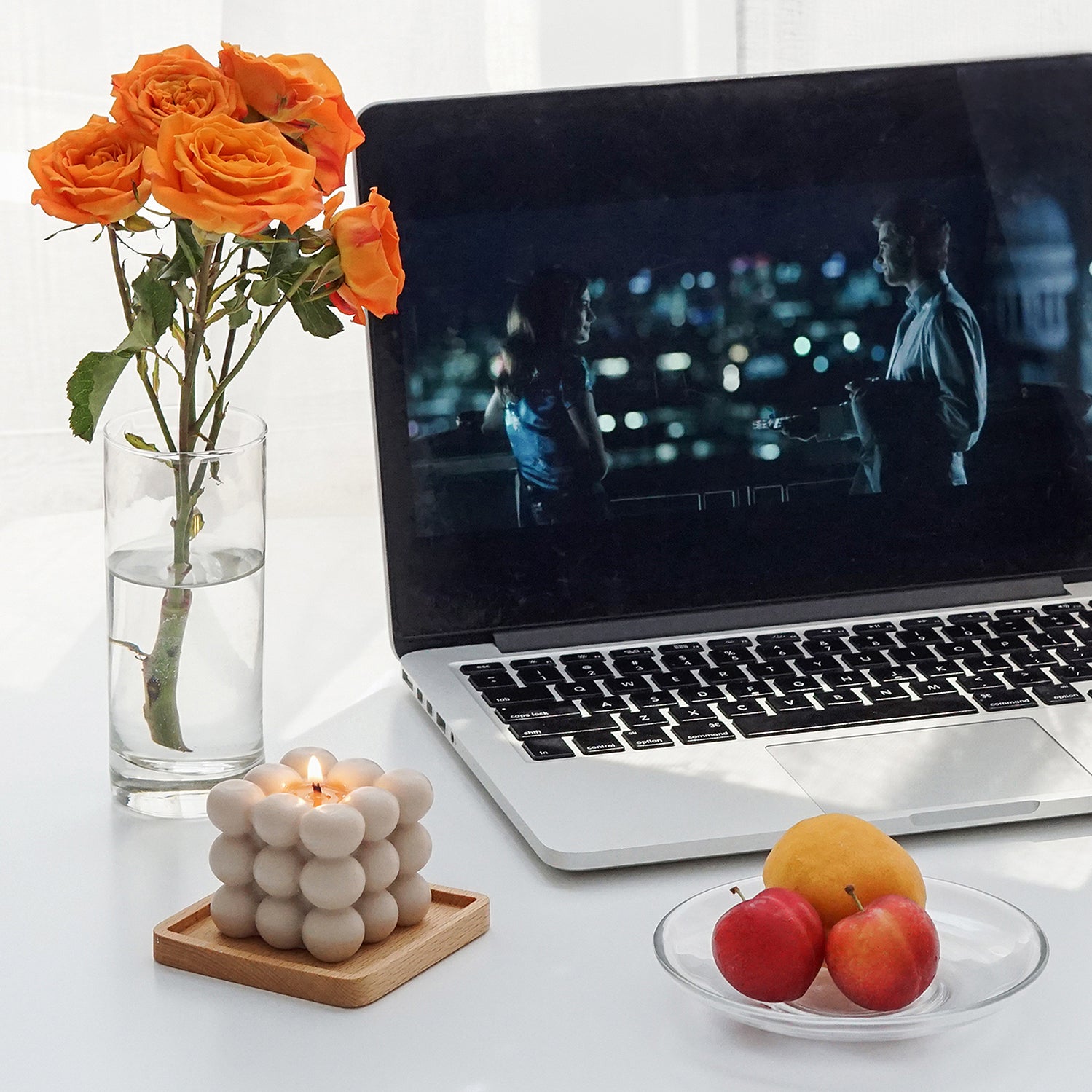 silver macbook displaying a movie scene from One Day, summer fruits on clear transparent dish, a lit beige square bubble cube soy pillar candle on a light wood square coaster, and orange roses in a cylinder clear transparent vase
