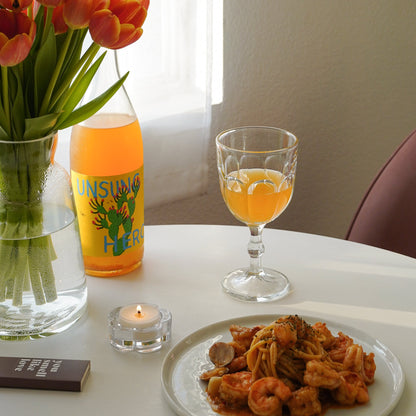 a lit tealight candle in a clear daisy candle holder, brown matchbox, pasta on a ceramic plate, a glass of orange wine, a bottle of orange wine, and tulips in a clear vase on white round table in an aesthetic room