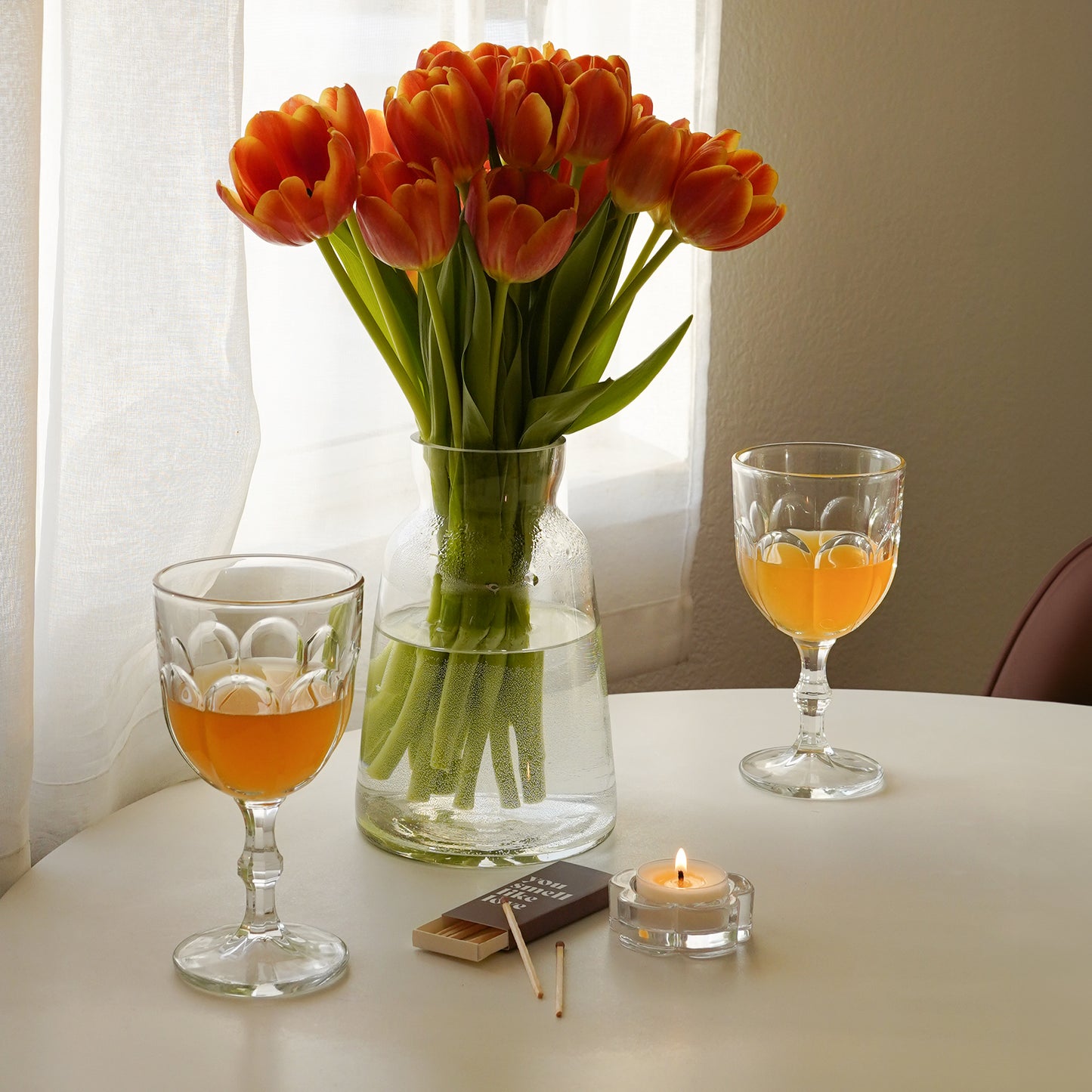 a lit tealight candle in a clear daisy candle holder, brown matchbox,  glasses of orange wine, and tulips in a clear vase on white round table in an aesthetic room